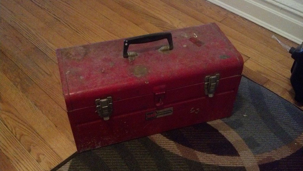A well used red metal toolbox resting on a rug and a wooden floor